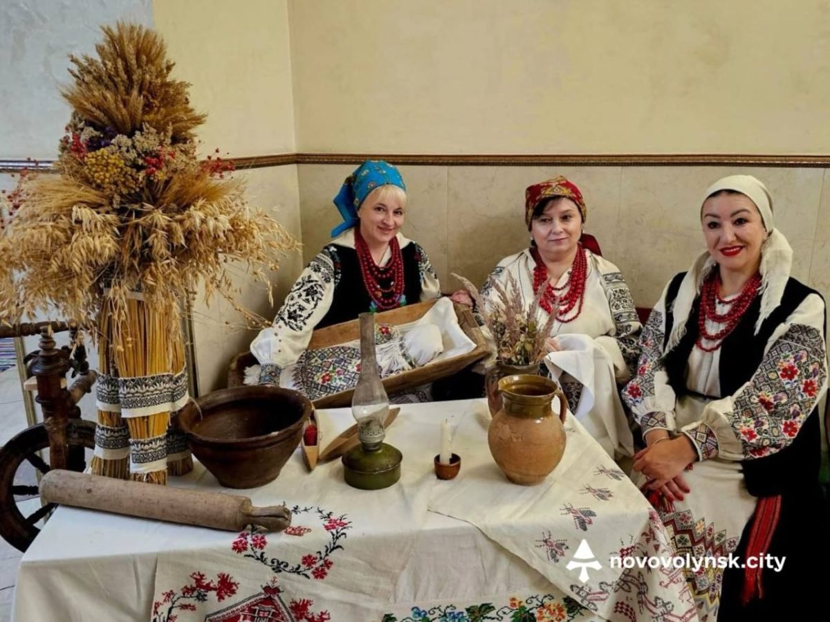 Три жінки в народних строях за столом з глечиками та гасовою лампою